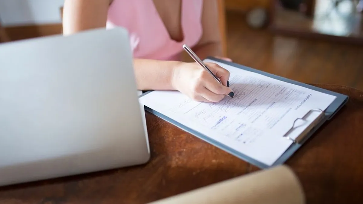 Person filling in a paper application form on a clipboard beside a laptop