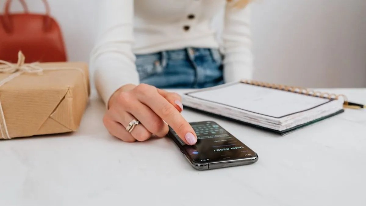 Hand using a smartphone beside a notebook and a wrapped parcel on a white desk