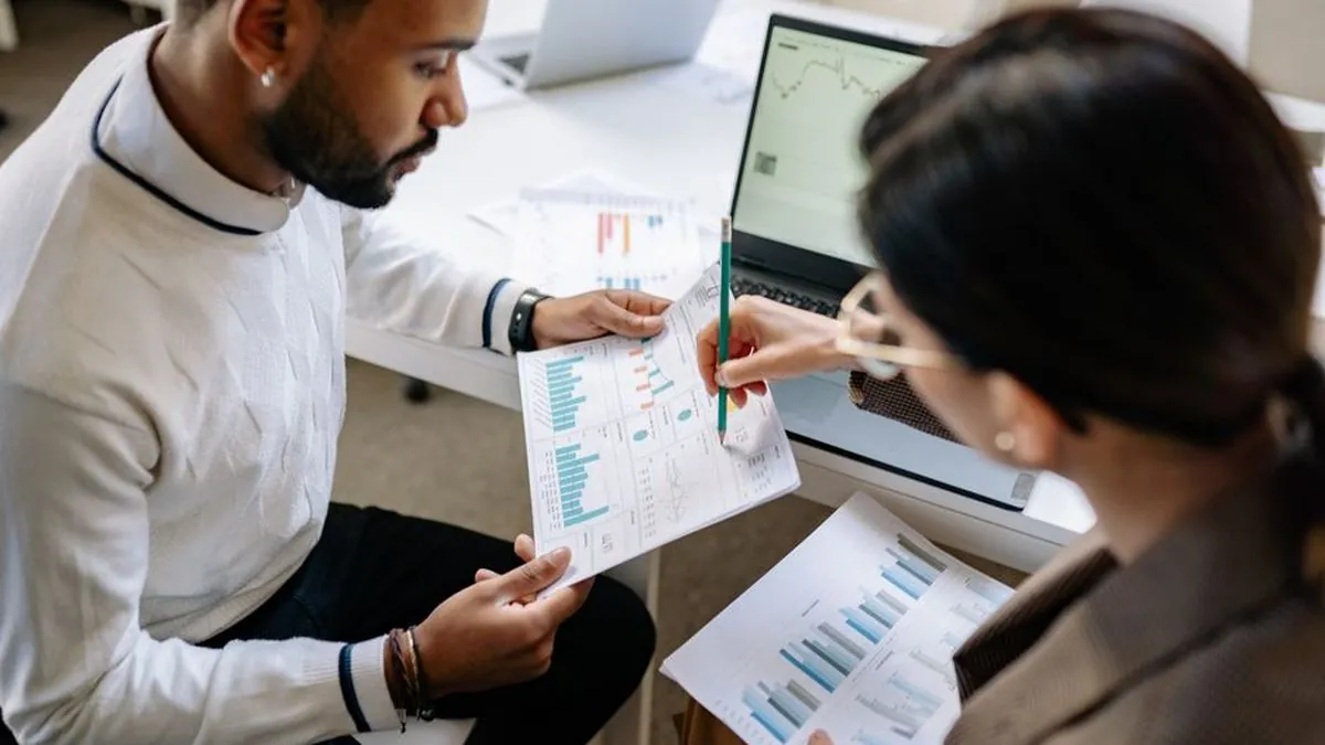 Two colleagues reviewing printed charts beside a laptop showing a performance graph