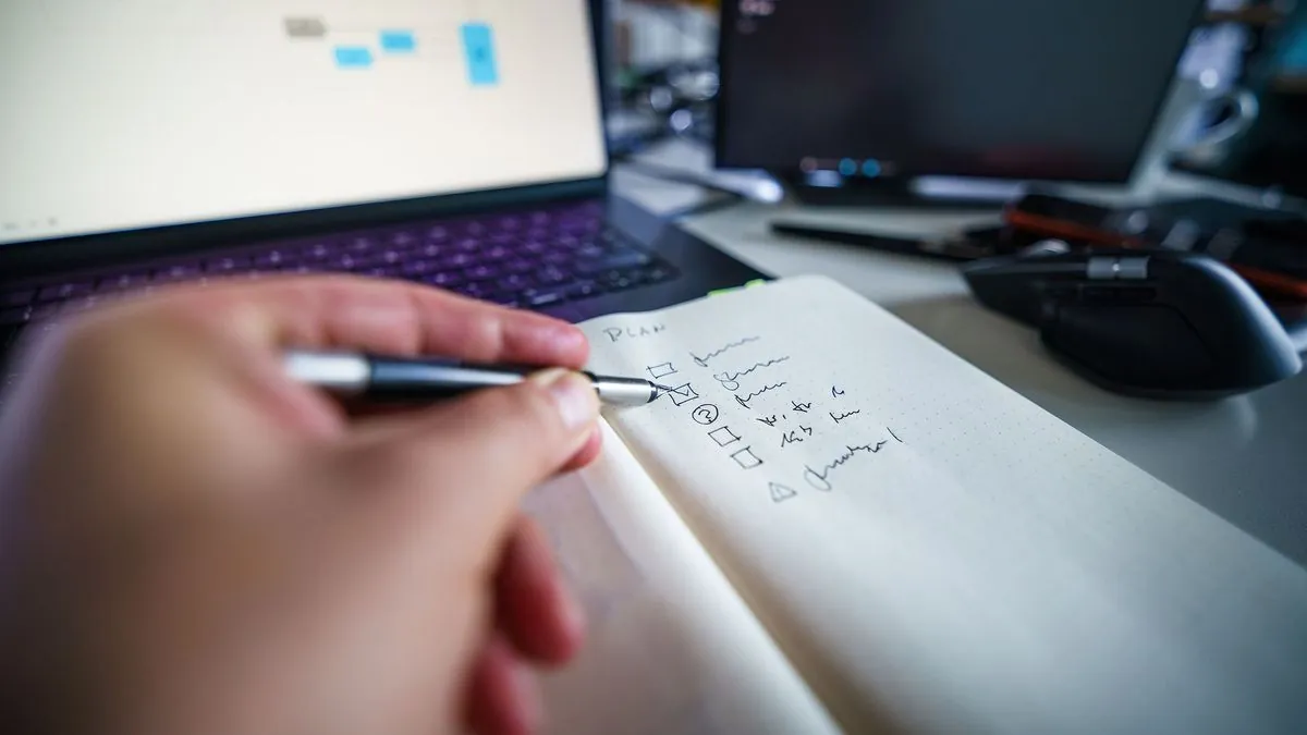 Hand ticking items on a plan checklist in a notebook beside a laptop