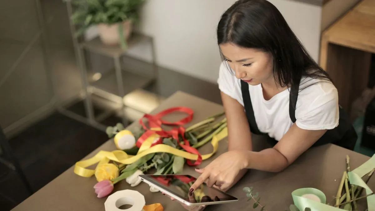 Florist checking a tablet at a work table covered in ribbons and flowers
