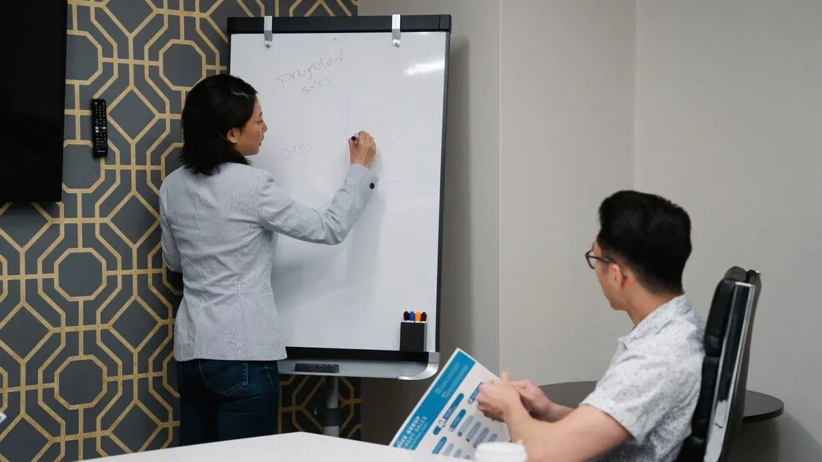 Woman writing on a whiteboard while a colleague sits at a table during a planning meeting