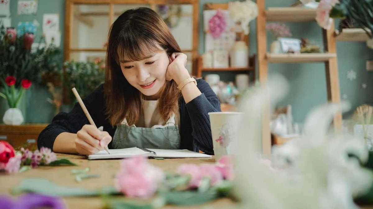 Smiling florist in an apron writing marketing notes in a notebook at her shop table