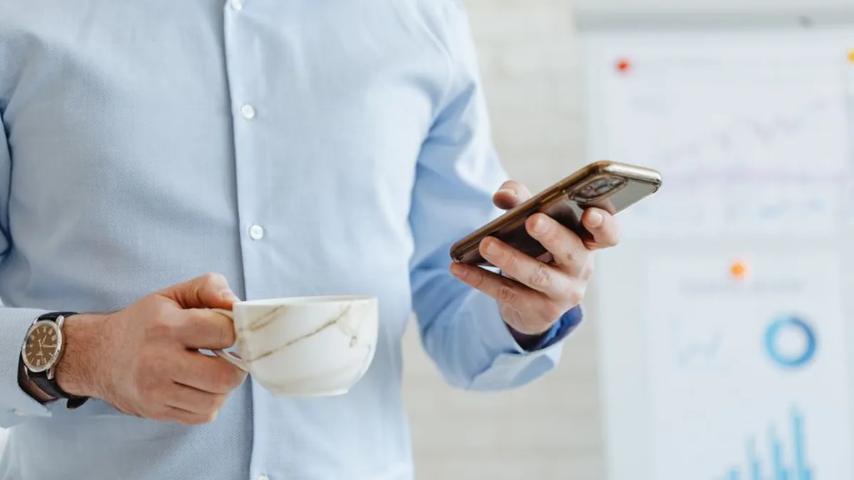 Man in a blue shirt holding a coffee cup and checking his smartphone