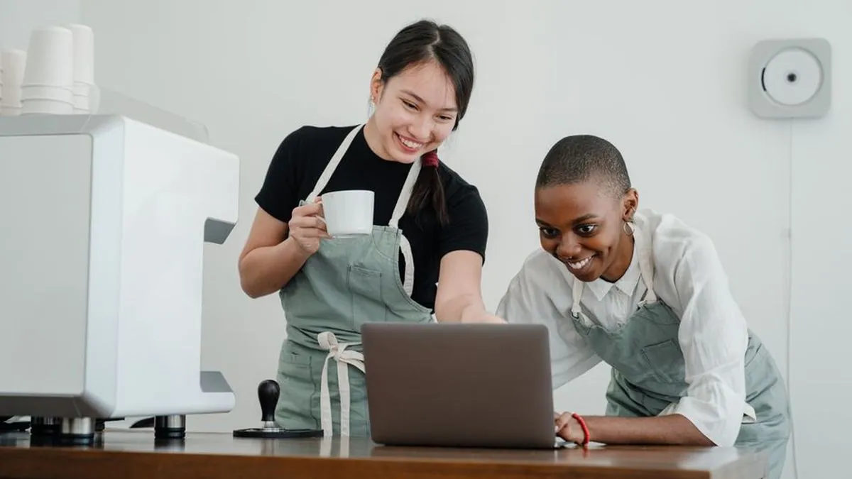 Two coffee shop workers smiling while looking at a laptop together