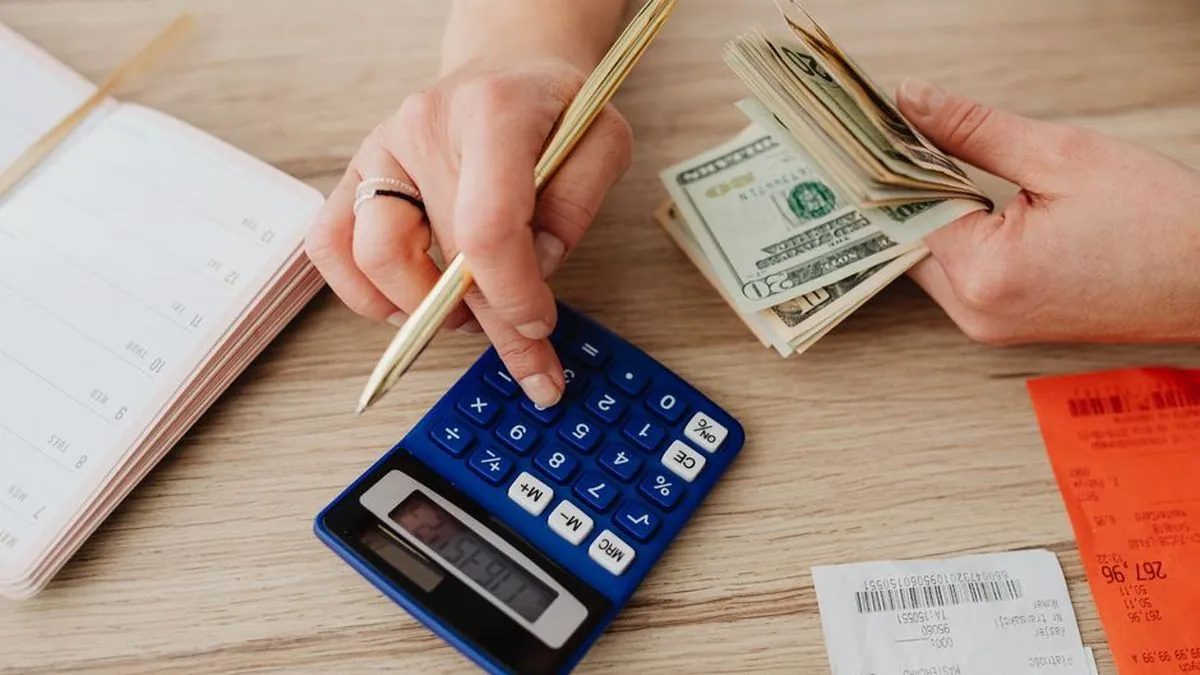 Hands using a calculator next to a notebook and cash on a wooden desk