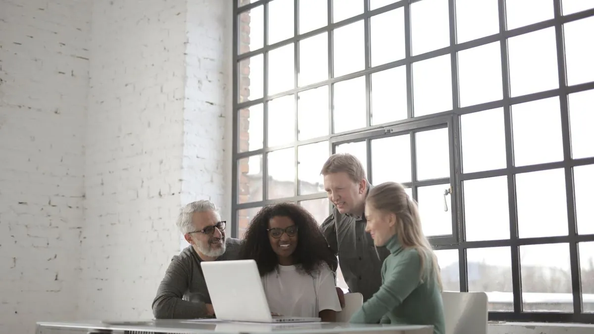 Four colleagues gathered around a laptop in a bright office with a brick wall and large windows