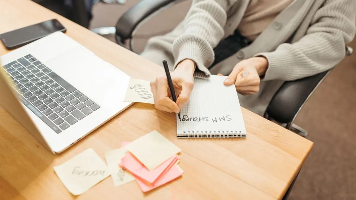 Person writing a plan in a notebook beside a laptop and sticky notes