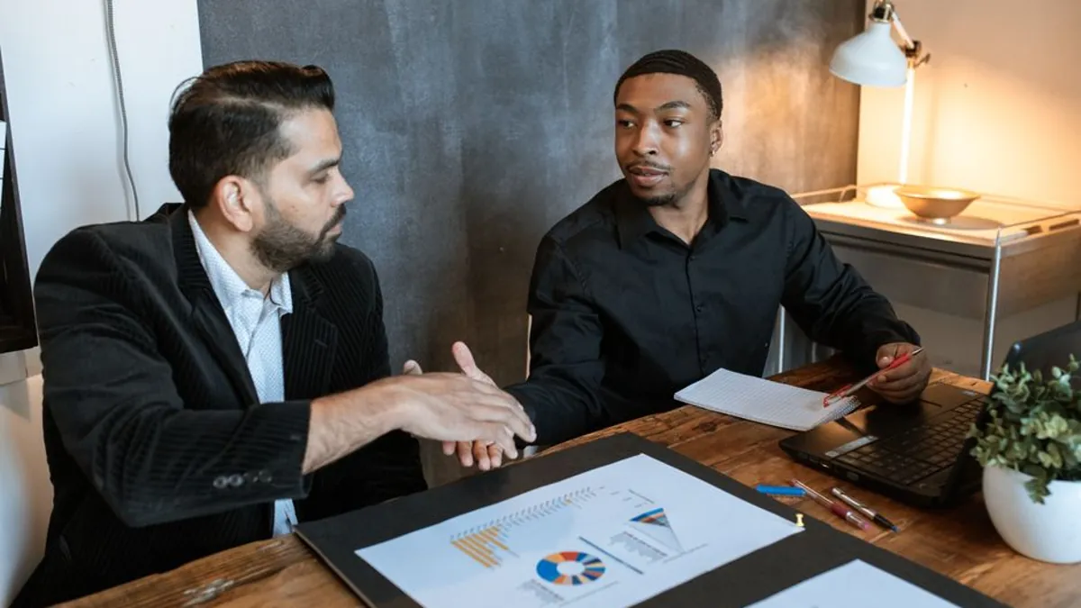 Two men shaking hands across a desk with printed charts between them