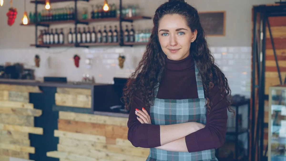 Smiling cafe owner in an apron standing with arms crossed at her counter
