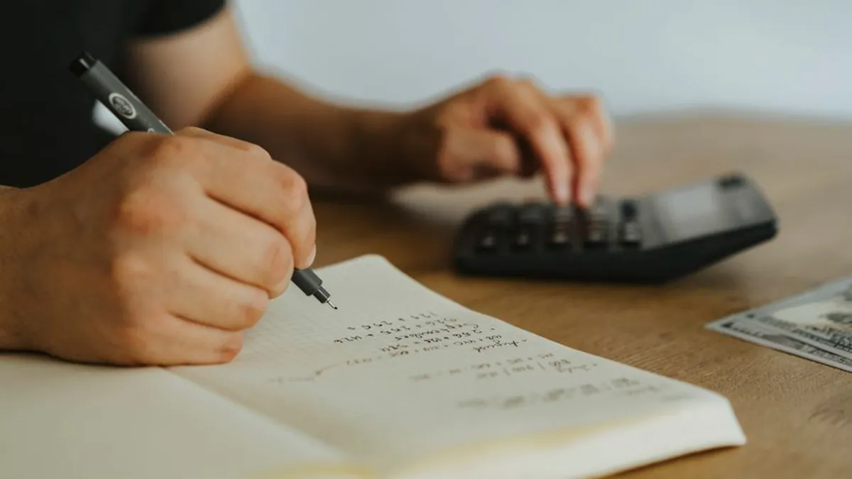 Hands writing a budget in a notebook beside a calculator on a wooden desk
