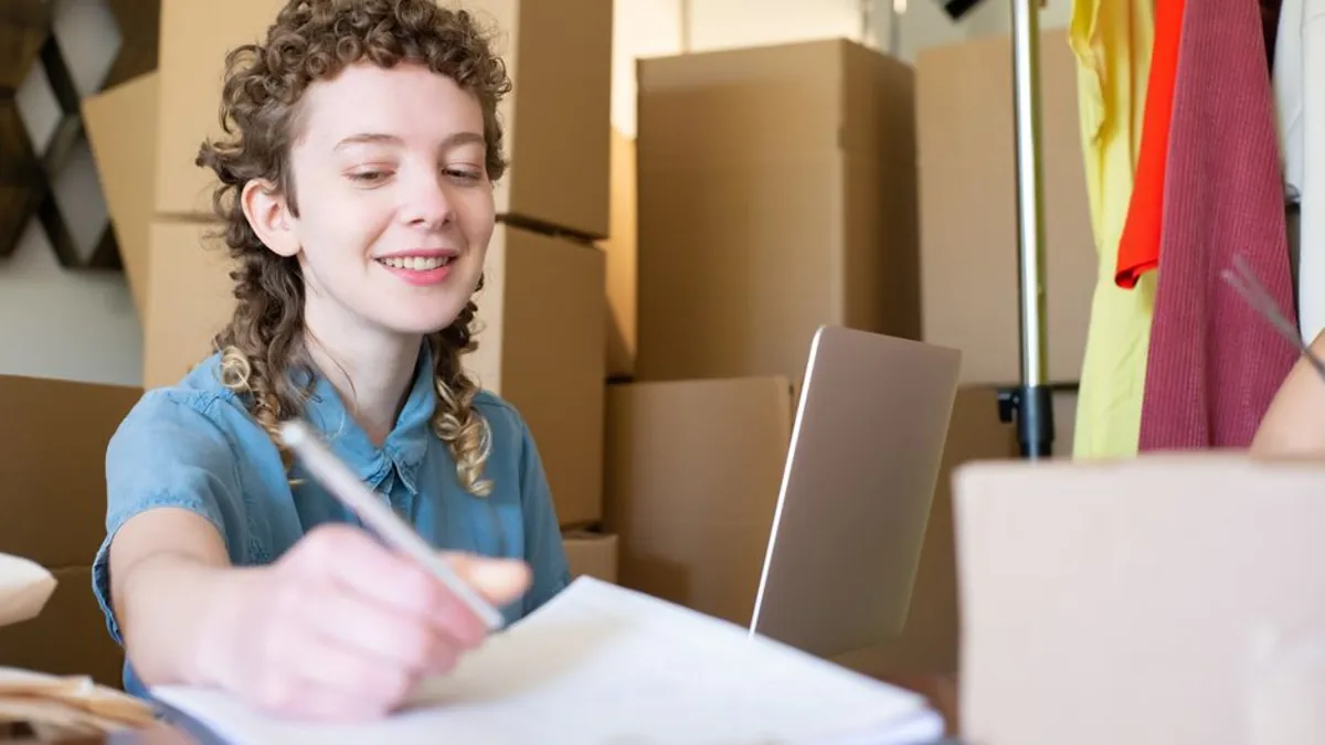 Woman writing notes in a notebook at a small online shop workspace with parcels