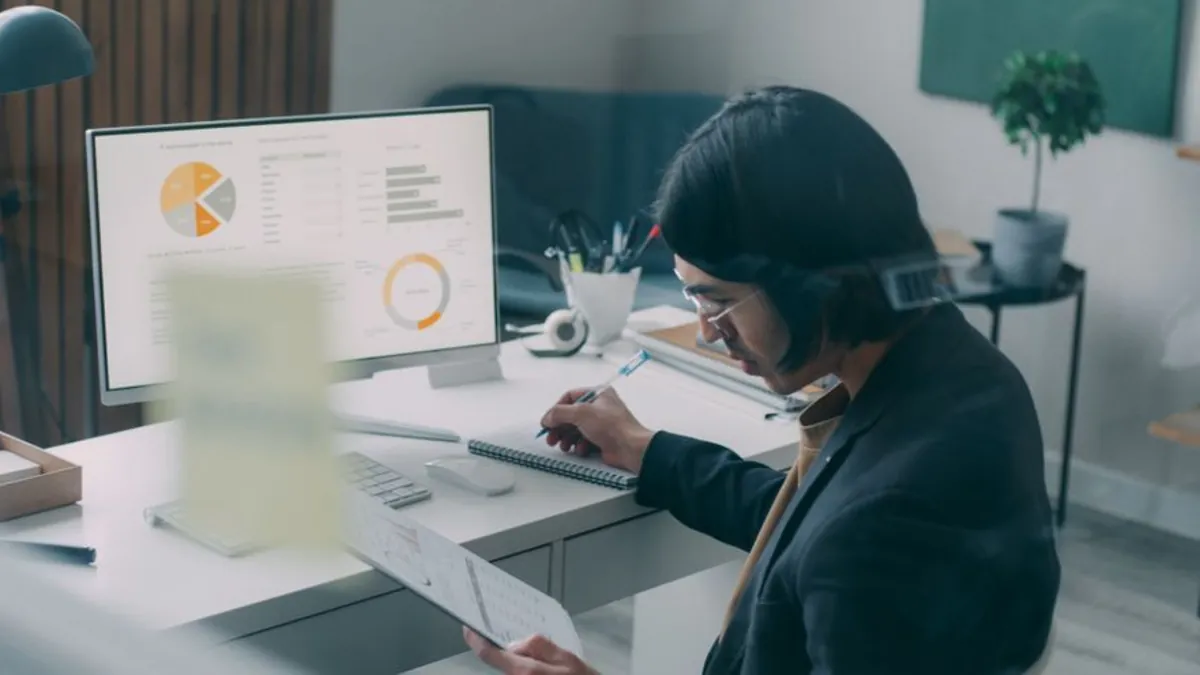 Analyst reviewing charts on a monitor and writing notes at a desk