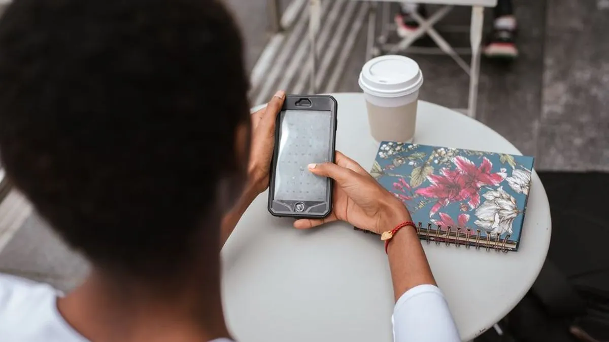 Person at a cafe table checking a smartphone beside a coffee cup and notebook